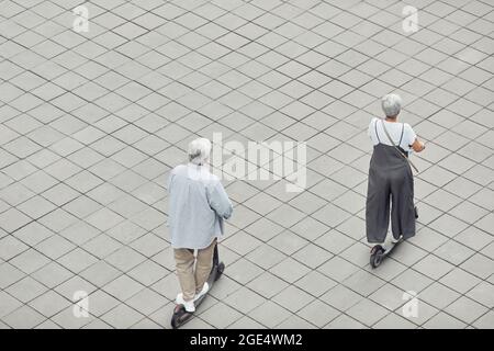 Back view at contemporary mature couple riding electric scooters in park across tiled background, copy space Stock Photo