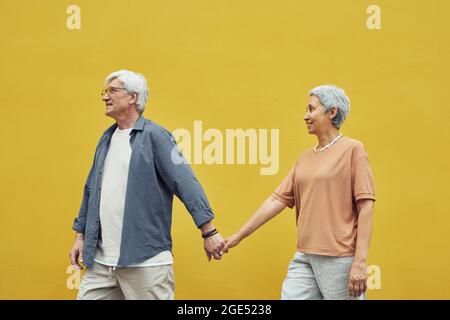 Pop color side view portrait of modern senior couple holding hands outdoors while walking against yellow background Stock Photo