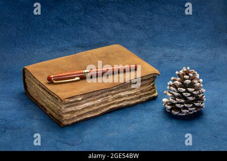 antique leather-bound journal with decked edge handmade paper pages with a stylish pen and a decorative pine cone against blue handmade paper, journal Stock Photo