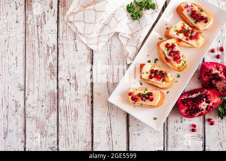 Holiday crostini appetizers with brie cheese, pomegranates and parsley. Above view table scene against a white wood background with copy space. Party Stock Photo