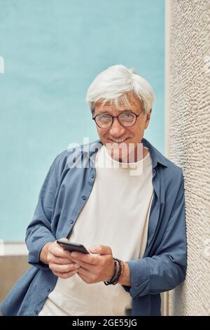 Vertical portrait of modern senior man using smartphone outdoors and smiling at camera while standing against light blue background Stock Photo