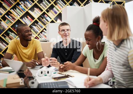 Diverse group of young people studying together at table in college library, focus on smiling young man chatting with friends Stock Photo
