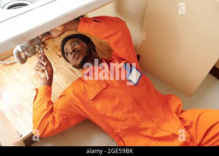 Young plumber in orange uniform lying under sink and fixing leaking pipe Stock Photo