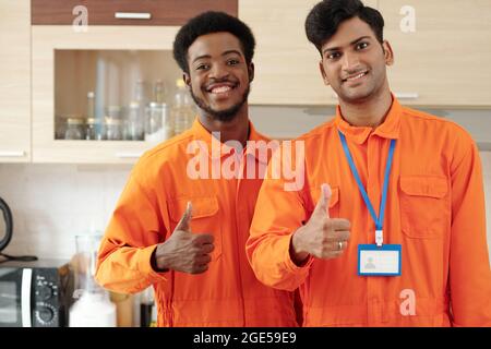 Team of positive plumbers in overalls smiling and showing thumbs-up when standing in kitchen Stock Photo