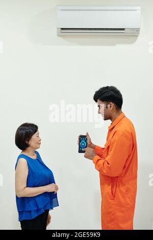 Technician explaining senior woman how to use air conditioner and change temperature in room via application on smartphone Stock Photo