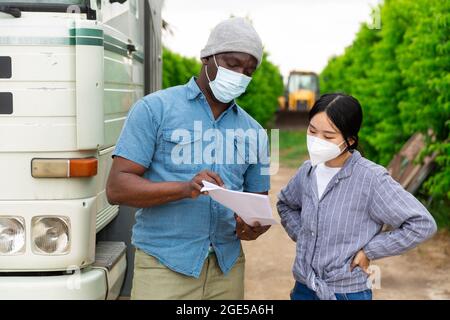 Two farm workers in face masks for disease protection discussing papers standing near car Stock Photo