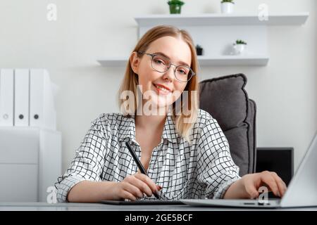 Young smiling woman draws on graphic tablet using laptop in office interior. Portrait Caucasian Blonde young happy woman in glasses freelance Stock Photo