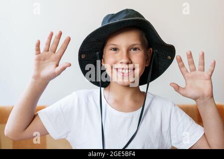 Cheerful school-age boy smiling while looking at the camera. In a white t-shirt and a black sun hat. Caucasian child having fun at the camera while sitting at home. Stock Photo