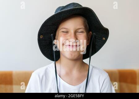 Cheerful school-age boy in white t-shirt and black hat sits on the couch and makes a funny grimace. Funny facial expression, close-up Stock Photo
