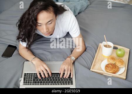 Attractive young man eating breakfast in bed and typing on laptop Stock Photo