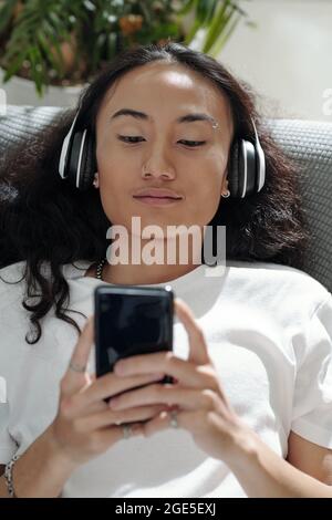 Portrait of smiling young man in headphones resting on sofa and watching funny videos on smartphone Stock Photo