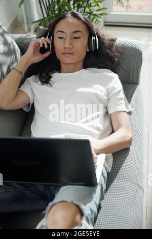Attractive young man lying on sofa, listening to music in headphones and programming on laptop Stock Photo