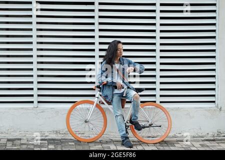 Fashionable young man in denim clothes leaning on bicycle and checking time or notifications on his smartwatch Stock Photo