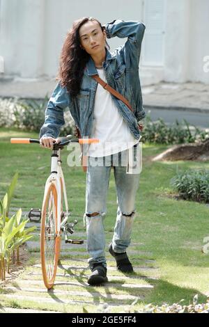 Full-length portrait of stylish young man in denim clothes standing at bicycle, touching his long curly hair and looking at camera Stock Photo