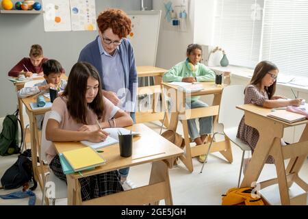 Clever schoolgirl writing down notes in copybook at lesson while her teacher standing near by Stock Photo
