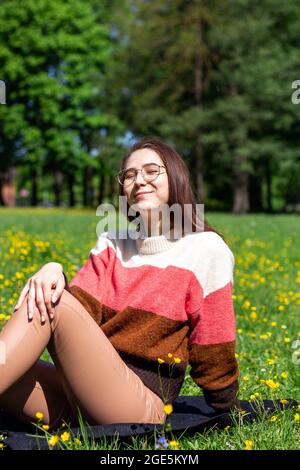 A beautiful young lady sitting on the grass enjoys life, feeling happy and relaxed in the park Stock Photo