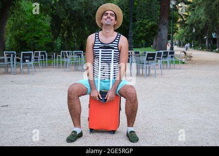 Tourist man smiling sitting on suitcase in a park. Stock Photo