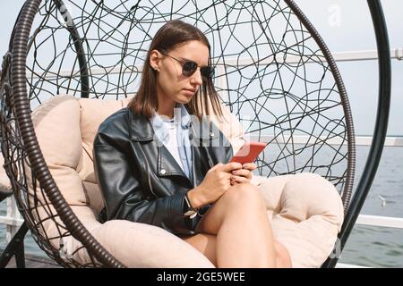 Serious young woman in sunglasses sitting in comfy chair outdoors and reading text messages from friends on smartphone Stock Photo