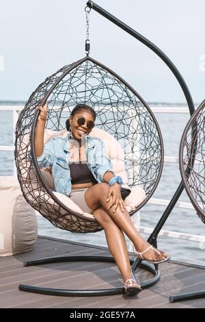 Smiling beautiful young woman dressed for outdoor party sitting in hanging chair and smiling at camera Stock Photo