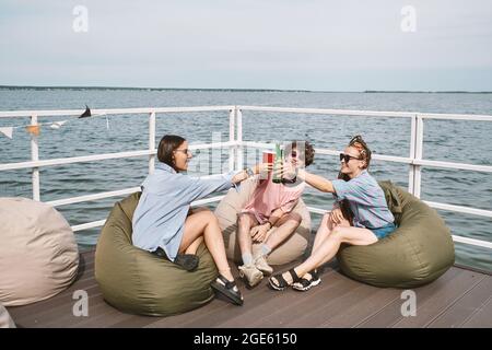 Group of happy young people sitting in bean bags on pier and toasting with beer bottles and plastic cups Stock Photo