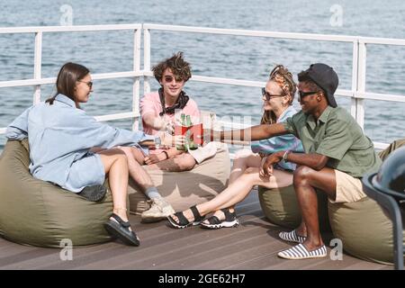 Smiling friends resting in bean bags on pier and toasting with plastics cups and beer bottles Stock Photo