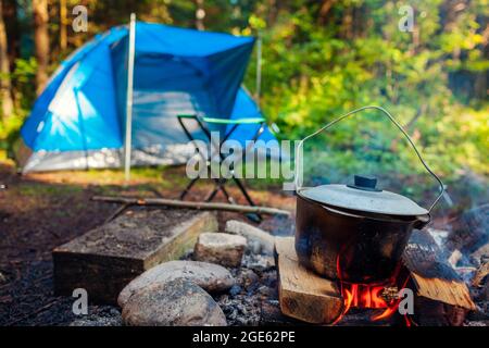 Camping in summer forest. Cooking on campfire in cauldron pot by hiking tent. Traveling and tourism Stock Photo