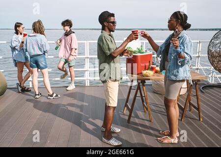Young people toasting with refreshing drinks when their friends talking in background at party on pier Stock Photo