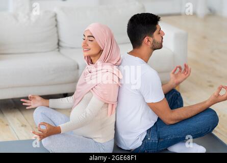 Yoga for pregnancy. Happy muslim spouses meditating together at home, pregnant arab woman in hijab sitting back to back with her husband, practicing l Stock Photo