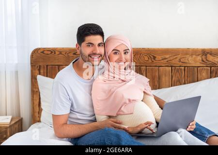 Loving pregnant muslim couple having fun at home, using laptop together, surfing internet and spending time together, sitting on bed and smiling at ca Stock Photo