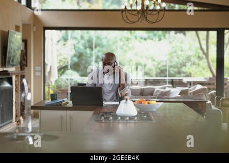 Senior african american man working on laptop and using smartphone in the modern kitchen Stock Photo