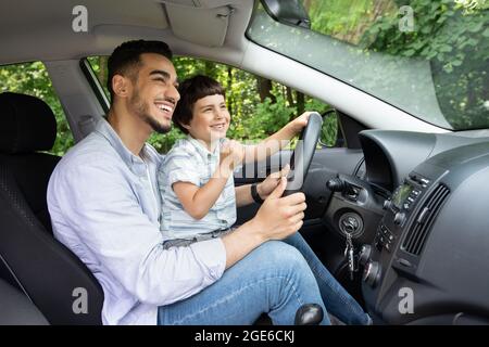 Young Arab Father Giving Driving Lessons To His Little Son, Happy Preteen Boy Sitting On Dad's Lap In Modern Car And Holding Steering Wheel, Enjoying Stock Photo