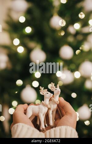 Hands holding reindeer ornament on background of christmas tree lights. Beautiful christmas deer toy in hands in festive decorated scandinavian room. Stock Photo