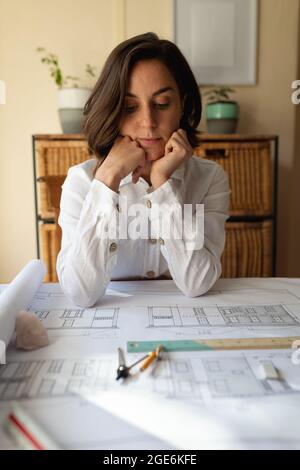 Caucasian female architect in living room, sitting at table working with plans Stock Photo