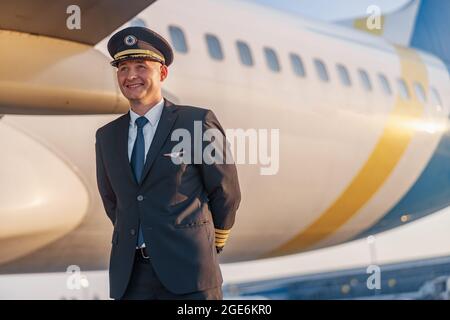 Portrait of smiling young male pilot in uniform and hat looking aside, posing in front of a big passenger airplane in airport at sunset Stock Photo