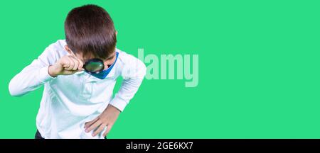 School age boy in a medical face mask blue color looking at an object with a magnifying glass holding in his hand. Stock Photo