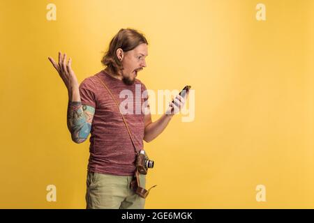 Portrait of young man, photographer, cameraman with retro camera using phone isolated on yellow studio background. Concept of occupation, job, funny Stock Photo