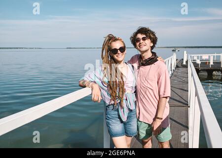 Hugging cheerful young boyfriend and girlfriend standing on pier, hugging and looking at camera Stock Photo