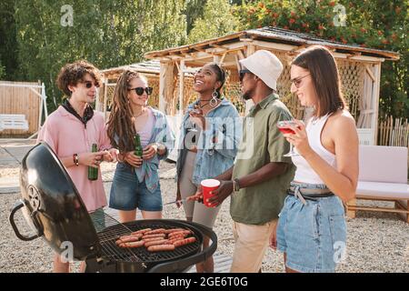 Group of friends drinking beer, grilling hot dogs and joking around at beach party on summer day Stock Photo