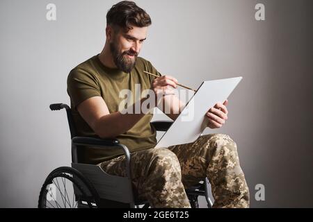 Military man sitting in a wheelchair and painting a picture with a brush Stock Photo