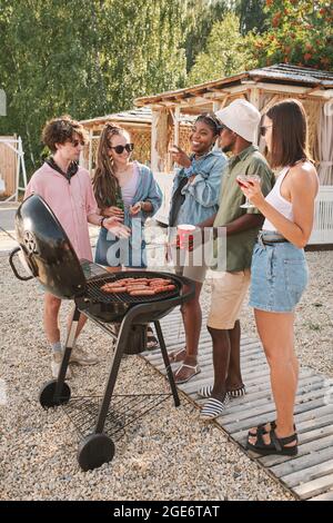 Pretty young woman telling joke to her friends at beach party when they are grilling hot dogs and drinking beverages Stock Photo