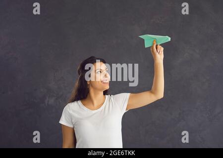 Portrait of a happy young woman holding a paper airplane in her hand standing on a gray background. Stock Photo