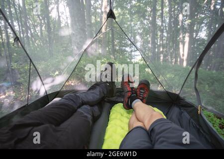 Male legs inside camp tent with summer forest on background. Two travelers lying inside tourist tent and enjoying the view of beautiful nature. Concept of travelling, hiking and camping Stock Photo