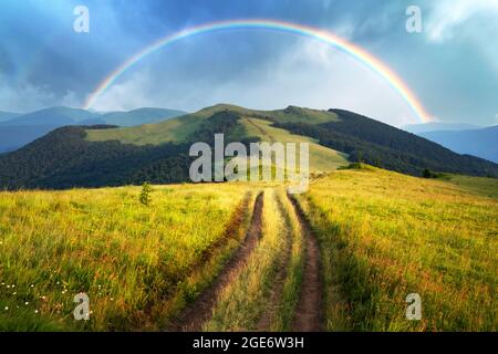Amazing scene in summer mountains. Lush green grassy meadows in fantastic evening sunlight. Rural road and beautyful rainbow in dramatic sky. Landscape photography Stock Photo