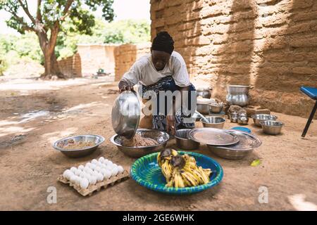 Black African housewife sitting in the middle of her cooking utensils, pouring boiled rice into a big metal dish Stock Photo