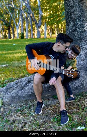 Brunette boy dressed casually and wearing sunglasses sitting in a tree playing classical guitar in the forest with his little son enjoying together as Stock Photo