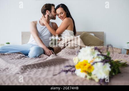 blurred bouquet of flowers near happy couple hugging on bed Stock Photo