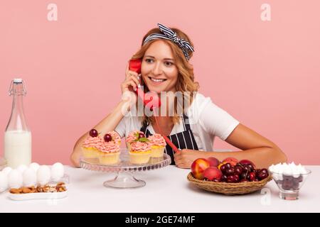 Portrait of smiling blonde young adult baker female talking via handset, talking about with customer, taking order, posing near ready-made cakes. Indo Stock Photo