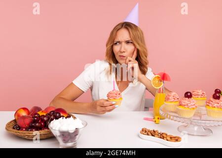Thoughtful blonde woman wearing party cone thinking about birthday desire, making wish, frowning face and keeps finger in cheek, holds cake with candl Stock Photo