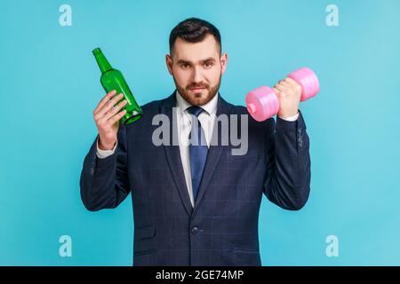 Portrait of bearded businessman in official style suit holding in hands beer bottle and pink dumbbell, choosing between alcohol and healthy lifestyle. Stock Photo