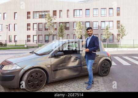 Portrait of young adult bearded insurance agent standing with positive facial expression near wrecked car and showing thumb up, wearing jeans and jack Stock Photo